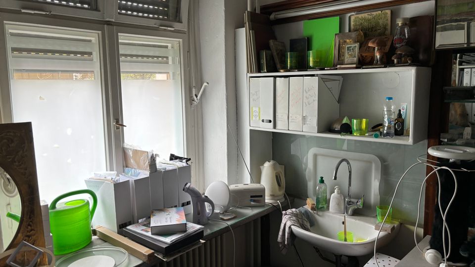 Kleiner, vollgestellter Arbeits- und Abstellbereich mit großen Fenstern und halb geschlossenen Rollläden, daneben ein Wandregal mit Ordnern und diversen Gegenständen. Rechts steht ein kleines Waschbecken mit Wasserhahn, Seifenspendern und Bechern, umgeben von Papierstapeln und Geräten auf den Arbeitsflächen.
