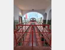 Innenansicht eines historischen Gefängnisses mit zwei Etagen und roten Metallgeländern. Die Zellen liegen entlang schmaler Gänge, und durch den Gewölbebogen ist eine zentrale, langgestreckte Halle mit heller Decke zu sehen.