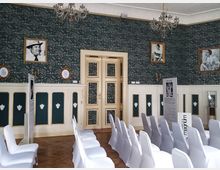 Innenraum eines historischen Veranstaltungsraums mit weiß eingedeckten Stühlen in Reihen, verzierten grünen Tapeten mit floralen Mustern und goldgerahmten Fotografien an den Wänden. Ein Kronleuchter und eine holzgetäfelte Wand mit Ornamenten prägen das Ambiente.