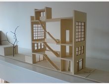 A minimalist architectural model of a multi-story building with open walls, featuring a series of staircases connecting the floors, large grid-style windows on each level, and a plain base with a small branch-like structure to the side.