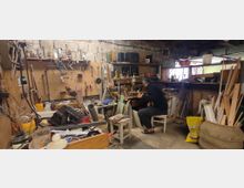 Eine unordentliche Werkstatt mit einer Vielzahl von Werkzeugen und Materialien, darunter Holzstücke, Rohre und Behälter, die auf Werkbänken und Regalen verteilt sind. Eine Person, die auf einem Hocker sitzt, arbeitet an einem Projekt in der Mitte des Raums.