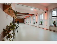 Heller, geräumiger Innenraum mit weißen Wänden, großen Fenstern an der rechten Seite und Holzelementen an der linken Wand, an der Pflanzen dekorativ angebracht sind. Der Raum hat einen glatten Boden und ist mit roten Lichtakzenten an den Wänden beleuchtet.