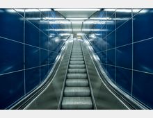 Ansicht einer Rolltreppe in einem modernen U-Bahn- oder Bahnhofsbereich mit glänzenden, dunkelblauen Kacheln an den Seitenwänden und hellem Deckenlicht. Die Rolltreppe führt nach oben in eine beleuchtete Passage.