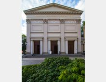 Fassade des Maxim Gorki Theaters in Berlin, ein klassizistisches Gebäude mit vier weißen Säulen, Verzierungen und einem Giebeldach. Vor dem Eingang befinden sich Stufen, Laternen und grüne Büsche im Vordergrund.