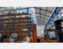 Innenansicht eines großen Lagerhauses mit hohen Regalen, die mit Kartons und Waren bestückt sind, teilweise hinter Gitter gesichert. Im Vordergrund befinden sich ein orangefarbener Gabelstapler sowie ein Schild mit der Aufschrift "Wareneingang", und die Decke zeigt ein Tragwerk aus blauen Stahlträgern mit Oberlichtern.