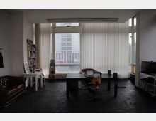 Ein minimalistisch eingerichtetes Büro mit einem großen Fenster mit vertikalen Jalousien, durch das ein Gebäude im Hintergrund sichtbar ist. Der Raum enthält einen schwarzen Schreibtisch mit einem Laptop, eine braune Ledercouch, ein Regal mit Büchern und eine weiße Wand.