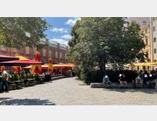 Ein belebter Platz mit Cafés und Sitzmöglichkeiten, flankiert von einem historischen roten Backsteingebäude mit verzierten Fenstern. Bunte Sonnenschirme spenden Schatten, während Menschen auf Bänken und unter Bäumen sitzen.