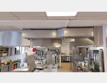 Große, funktionale Großküche mit Edelstahl-Arbeitsflächen, industriellen Dunstabzugshauben und mehreren Küchenmaschinen. Im Hintergrund sind Regale mit Geschirr, Töpfen und anderen Küchenutensilien sowie Fliesenwände sichtbar.