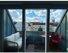 Ein kleiner Balkon mit Glasbrüstung, ausgestattet mit einem weißen Sofa, einem roten Tisch und zwei roten Stühlen. Im Hintergrund sind mehrstöckige Wohnhäuser mit Satteldächern und ein Kirchturm vor einem Himmel mit Wolken zu sehen.