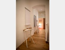 Schmaler Innenraum mit Holzboden und heller Wandgestaltung, ausgestattet mit einem Flipchart, kleinen Tischen und weißen Stühlen. Im Hintergrund ist ein Bogen, der zu einem beleuchteten Bereich mit einer Stehlampe führt.