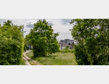 Ein schmaler Fußweg führt durch eine Wiese, die von hohen grünen Bäumen und Sträuchern gesäumt ist. Im Hintergrund sind moderne Wohngebäude und ein weißes Haus zu sehen, unter einem bewölkten Himmel.