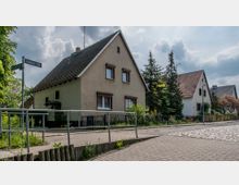 Straßenszene in einem Wohngebiet mit freistehenden Einfamilienhäusern, roten Satteldächern und cremefarbenen Fassaden. Im Vordergrund eine Gehwegkante mit Pflanzen und ein Straßenschild mit der Aufschrift „Fichtestraße“, umgeben von Bäumen und Gärten.