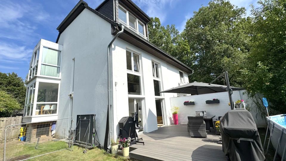 Modernes, weiß verputztes Einfamilienhaus mit dunklem Satteldach, großen Fensterflächen und einem gläsernen Anbau an der Seite. Davor liegt eine Holzterrasse mit Gartenmöbeln, Sonnenschirm und Grill, umgeben von Rasen und dichtem Baumbestand.