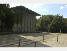 Außenansicht eines Gebäudes aus Beton mit einer flachen, vorstehenden Dachkonstruktion und bewachsenen Wänden. Der Vorplatz ist mit Steinplatten in einem Schachbrettmuster gestaltet, umgeben von Bäumen und Grünflächen.