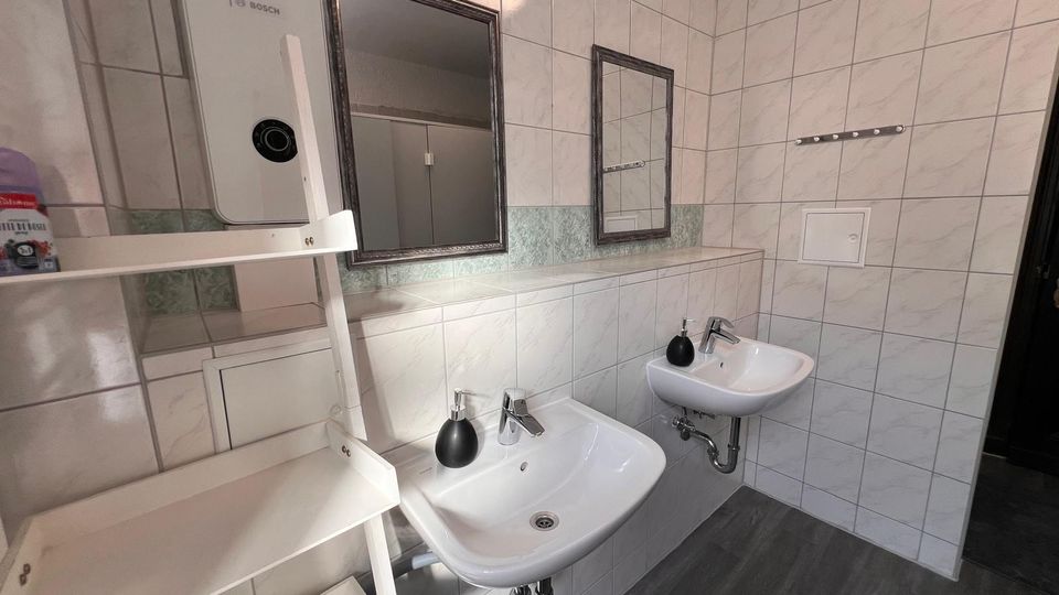 Gefliester Waschraum mit zwei wandmontierten Waschbecken, Seifenspendern und zwei Spiegeln über einer langen Ablage. Links steht ein offenes Regal mit einem wandhängenden Warmwassergerät; der Boden ist dunkel und wirkt wie Vinyl oder Laminat.