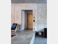 Modernes Wohnzimmer mit Betonwänden und -boden, einer Holztür in einem Wanddurchbruch und schlichter Einrichtung, darunter ein hölzerner Sessel mit dunkelblauem Polster und ein kleiner Stapel Zeitschriften in einer Halterung.