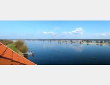 Weitläufige Flusslandschaft mit ruhiger Wasseroberfläche, umgeben von vereinzelten Bäumen und sandigen Uferbereichen; links unten ein Abschnitt eines roten Ziegeldaches und ein kleiner Bootsanleger. Der Himmel ist klar mit einigen weißen Wolken.