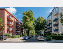 Straßenansicht einer Wohngegend mit mehrstöckigen Gebäuden auf beiden Seiten, die Balkone mit blauen Geländern haben. In der Mitte verläuft eine von Bäumen gesäumte Kopfsteinpflasterstraße, an der Autos geparkt sind.