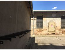 Ein Industriehof mit alten, verwitterten Gebäuden. Die Wand links hat eine rostige Metallhalterung mit Ketten, während die gegenüberliegende Wand Fenster mit Gitterrahmen und eine verschlossene Metalltür zeigt; der Boden besteht aus Beton.