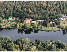Luftaufnahme eines Flussufers mit mehreren Häusern und Gärten, die entlang des Wassers verlaufen. Im Hintergrund ist ein dichter Wald zu sehen, der das Gebiet umrahmt.