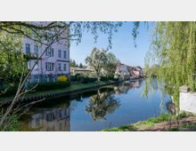 Ein ruhiger Fluss, gesäumt von grünen Böschungen und Bäumen, die ins Wasser hängen, mit Wohnhäusern im Hintergrund, darunter ein mehrstöckiges Gebäude mit pastellfarbener Fassade. Die Umgebung ist von Gartenanlagen, blühenden Bäumen und klar reflektierendem Wasser geprägt.