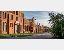 Ziegelrote Industriegebäude mit großen Rundbogenfenstern, aufgereiht entlang einer gepflasterten Straße mit Straßenbahnspuren; daneben Grünflächen und ein Baum im Vordergrund.