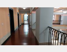 Ein Innenbereich eines Gebäudes mit einem Flur, orangefarbenen Wänden und schwarzen Türrahmen. Der Flur führt zu einem angrenzenden Bereich, während rechts ein Geländer mit schwarzen Metallstreben eine Öffnung zu einem darunterliegenden Stockwerk abgrenzt.