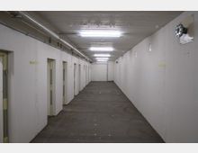 Ein schlichter, langgestreckter Korridor mit weißen Betonwänden und mehreren Türen auf der linken Seite. Die Decke ist mit sichtbaren Rohren und mehreren Leuchtstofflampen ausgestattet; der Boden besteht aus grauem Beton.