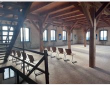 Großer Raum mit sichtbaren Holzbalken und unverputzten Wänden, ausgestattet mit einer Reihe moderner Holzstühle. Große Fenster lassen viel Tageslicht herein, links führt eine Holztreppe ins Untergeschoss.