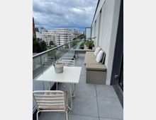 Moderne Dachterrasse mit Glasbrüstung und grauem Fliesenboden, ausgestattet mit einem weißen Tisch, zwei weißen Stühlen und einer gepolsterten Sitzbank. Im Hintergrund eine Stadtansicht mit modernen Gebäuden, einer Kirche aus rotem Backstein und bewölktem Himmel.