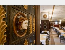 Detailansicht eines historischen Innenraums mit verzierten Wandreliefs, die ein Gesichtsmotiv zeigen, im Vordergrund. Im Hintergrund ein großzügiger Gastraum mit gedeckten Tischen, holzvertäfelten Wänden und hoher Decke mit schlichten Verzierungen.