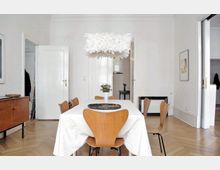 Esszimmer in einem minimalistischen Stil mit einem rechteckigen Tisch, der mit einer weißen Tischdecke bedeckt ist, und sechs Holzstühlen. Eine große, weiße Federlampe hängt von der Decke, dahinter sind weiße Türen und helle Wände zu sehen, ergänzt durch einen Holzboden mit Fischgrätmuster.