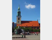 Historische Kirche mit rotem Ziegeldach und grünem Kirchturm, vermutlich die Marienkirche in Berlin. Im Vordergrund steht der Neptunbrunnen mit Figuren und Wasserspielen, umgeben von Bäumen und Menschen auf einem öffentlichen Platz bei klarem Himmel.