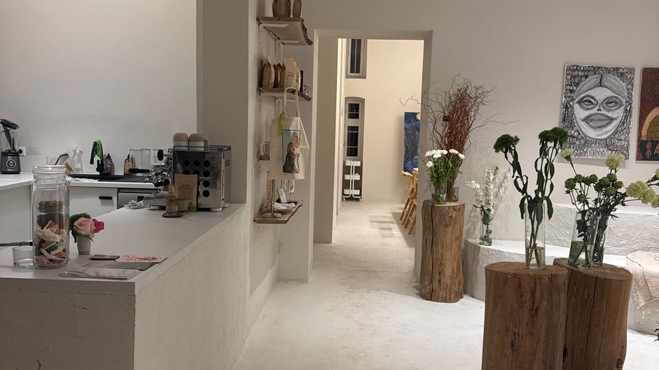 Helles, minimalistisches Innenraumstudio mit weißem Boden und Wänden, links eine Theke mit Kaffeemaschine und Regalen. Rechts stehen Baumstamm-Sockel mit Vasen und Blumenarrangements, daneben Sitzpolster und Wandkunst; ein Durchgang führt in einen hinteren Raum.