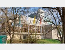 Modernes Gebäude mit großen Glasfenstern und hellem Stein, umgeben von Bäumen. Mehrere bunte Fahnen sind auf Masten vor dem Gebäude angebracht, und es befindet sich in einer grünen Parklandschaft.