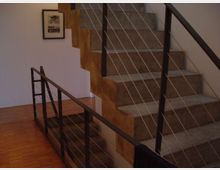 Treppe in einem modernen Innenraum mit Betonstufen und einem Geländer aus Metallstreben und dünnen Drahtseilen, auf einem Boden aus Holzlaminat; an der weißen Wand hängt ein gerahmtes Bild.