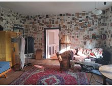 Wohnzimmer mit Vintage-Charakter: Wände mit zahlreichen Zeitungsausschnitten bedeckt, ein gelber Kachelofen links, ein roter Orientteppich auf Holzdielenboden, klassische Möbel wie ein lila Sofa, ein Retro-Lampenschirm und ein Holztisch. Ein Kleiderständer und ein Regal mit Büchern ergänzen die Einrichtung.