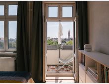Blick aus einem Zimmer mit hellen Wänden und dunklen Vorhängen durch eine geöffnete Balkontür. Auf dem Balkon befinden sich ein weißer Liegestuhl und Fliesen mit Blumenmustern, im Hintergrund ist eine Kirche mit spitzem Turm und umliegenden Gebäuden sichtbar.