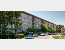 Wohnstraße mit einem mehrstöckigen Wohnblock im Hintergrund, der eine Fassade in Braun- und Grautönen aufweist. Entlang der Straße parken Autos, und Bäume sowie ein Verkehrsschild für eine Tempo-30-Zone sind sichtbar.