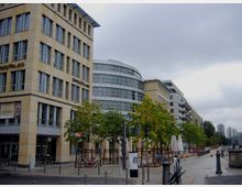 Moderne Gebäudezeile an einem städtischen Uferbereich, mit einem Eckgebäude namens "Spreepalais". Vor den Gebäuden stehen Bäume und Sitzbereiche mit Sonnenschirmen, im Hintergrund ist eine Promenade sichtbar.