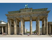 Das Bild zeigt das Brandenburger Tor in Berlin, ein monumentales Tor mit sechs dorischen Säulen und einer Quadriga-Statue auf der Spitze. Vor dem Tor versammeln sich mehrere Menschen auf einem gepflasterten Platz, und im Hintergrund sind moderne Gebäude sowie der Berliner Fernsehturm sichtbar.