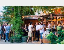 Eine belebte Outdoor-Bar mit Holzständen, Tischen und Bänken, umgeben von Pflanzen und Lichterketten. Menschen stehen und sitzen in Gruppen, plaudern und genießen die entspannte Atmosphäre unter Bäumen.