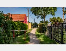 Schmaler, unbefestigter Weg mit Rasenstreifen, flankiert von Hecken, einem Zaun und einem Baum. Links steht ein rotes Haus mit Ziegeldach, im Hintergrund ein gelbes Gebäude; Stromleitungen verlaufen über den Weg.