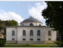 Gebäude mit klassizistischer Architektur, charakterisiert durch einen weißen Rundbau mit einer großen Kuppel und hohen, bogenförmigen Fenstern. Das Gebäude steht in einer parkähnlichen Umgebung mit Bäumen und Rasenflächen.