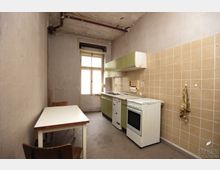 Schmale, einfache Küche mit einem abgenutzten Erscheinungsbild, bestehend aus einer Arbeitszeile mit grünen Oberschränken, einem weißen Herd und einem kleinen Tisch mit zwei Holzstühlen. Die Wände sind teils mit beigen Fliesen, teils unverputzt, und die Decke zeigt sichtbare Abnutzung, während ein Fenster am Ende des Raumes Tageslicht hereinlässt.