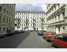 Innenhof mit Kopfsteinpflaster, umgeben von hellen, mehrstöckigen Altbauten mit verzierten Fassaden und Balkonen; parkende Autos säumen die Straße unter einem teils bewölkten Himmel.