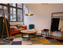 Ein gemütlicher Raum mit einem orangefarbenen Ledersofa, einem runden Holztisch und einer Stehlampe mit goldener Leuchte. Im Hintergrund sind große Fenster mit Blick auf eine Backsteinfassade, und an der Seite stehen Kleiderständer mit Mänteln sowie ein Hocker mit gefalteter Kleidung. Der Boden ist mit einem bunten Teppich in geometrischem Muster ausgelegt.