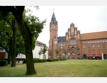 Backstein-Gebäude mit gotischen Elementen und einem hohen Turm mit Uhr, umgeben von Bäumen und einer Rasenfläche im Vordergrund; rechts neben dem Gebäude sind Sonnenschirme aufgestellt, die zu einem Café oder Restaurant gehören.