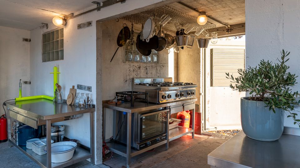 Eine einfache Außenküche mit Betonwänden, Edelstahlarbeitsflächen und einem Gasherd mit Backofen. Über der Küche hängen Kochutensilien, und seitlich ist eine Pflanze in einem grauen Topf sichtbar.