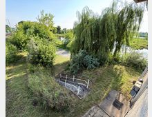 Grüne Landschaft mit einem Weg und einer Metallgeländetreppe, umgeben von Gras, Sträuchern und einem großen hängenden Baum (Weide). Im Hintergrund sind Bäume, ein kleiner See und ein Gehweg zu sehen.